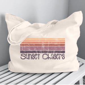 May include: Cream-colored tote bag with the text "Sunset Chasers" in a distressed font. The design features horizontal stripes in shades of orange, brown, and purple, evoking a sunset theme. The bag is made of a soft, durable fabric.