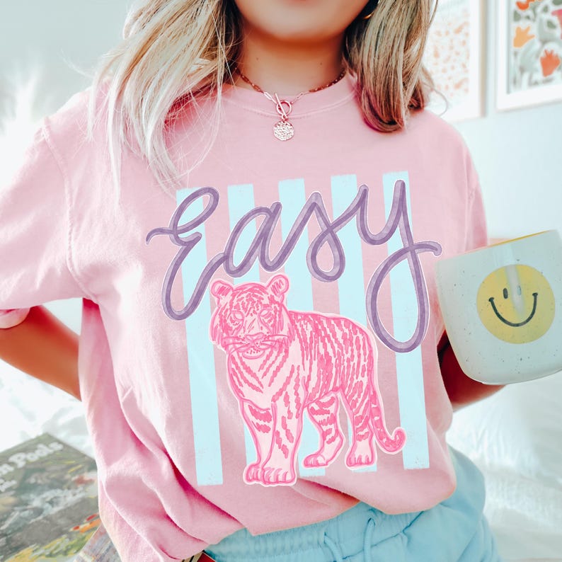 K&ouml;nnte beinhalten: Hellrosa T-Shirt mit dem Wort "Easy" in Schreibschrift und einem rosa Tiger-Motiv. Hinter dem Text und dem Tiger befinden sich hellblaue vertikale Streifen. Eine wei&szlig;e Tasse mit einem gelben Smiley wird in der Hand gehalten.