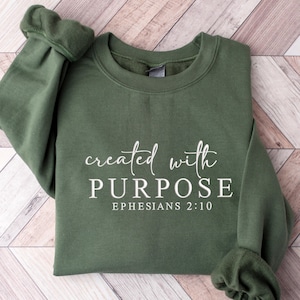 May include: An olive green sweatshirt featuring the white text "created with PURPOSE" in a script font, with "EPHESIANS 2:10" below in a serif font. The sleeves are visible. The garment is folded.