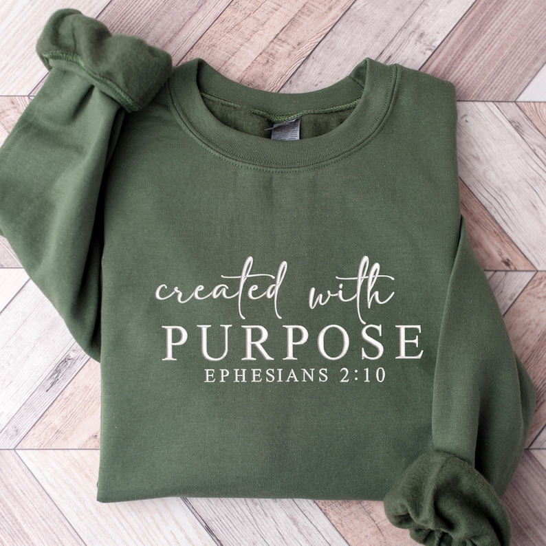 May include: An olive green sweatshirt featuring the white text "created with PURPOSE" in a script font, with "EPHESIANS 2:10" below in a serif font. The sleeves are visible. The garment is folded.
