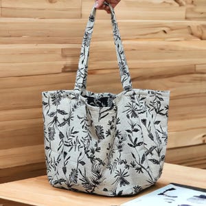 May include: A large, beige tote bag with black floral print. The bag has two long straps and a wide opening.