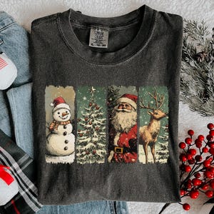 May include: Dark gray Comfort Colors t-shirt featuring a Christmas-themed graphic. The graphic includes a snowman, Christmas tree, Santa Claus, and a reindeer. The shirt has a tag that reads "XL - TG - EG".