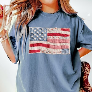 K&ouml;nnte beinhalten: Ein blaues T-Shirt mit einem Patchwork-Design der amerikanischen Flagge. Die Flagge zeigt rote und wei&szlig;e Streifen sowie ein Feld wei&szlig;er Sterne auf blauem Grund. Die Flagge besteht aus verschiedenen gemusterten Stoffen.