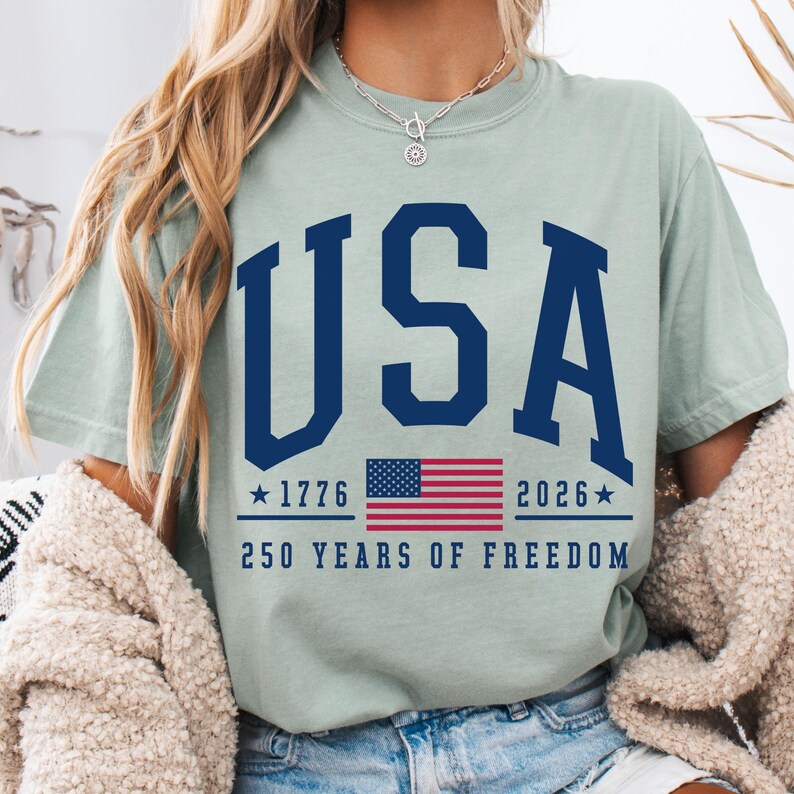 May include: A light green t-shirt with the word "USA" in navy blue block letters. Below the word "USA" is a graphic of the American flag and the text "1776" and "2026" with stars. The text "250 YEARS OF FREEDOM" is below the flag.
