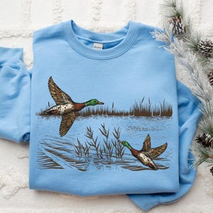 K&ouml;nnte beinhalten: Hellblaues Sweatshirt mit detaillierter Illustration von zwei Enten im Flug &uuml;ber Wasser und Schilf. Die Enten haben braune und wei&szlig;e Fl&uuml;gel und gr&uuml;ne K&ouml;pfe. Das Design ist vor dem Hintergrund einer Wasserszene.