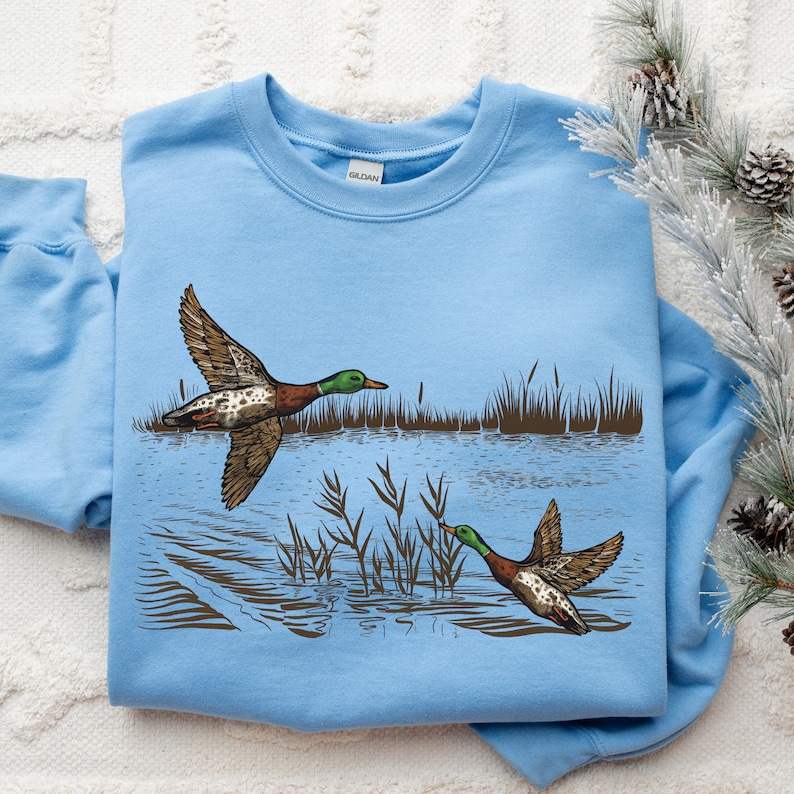 K&ouml;nnte beinhalten: Hellblaues Sweatshirt mit detaillierter Illustration von zwei Enten im Flug &uuml;ber Wasser und Schilf. Die Enten haben braune und wei&szlig;e Fl&uuml;gel und gr&uuml;ne K&ouml;pfe. Das Design ist vor dem Hintergrund einer Wasserszene.