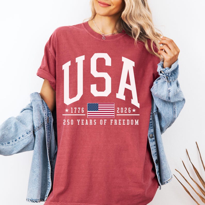 May include: A maroon t-shirt with the word "USA" in large white block letters. Below the word "USA" is a graphic of the American flag, with the years "1776" and "2026" on either side. The text "250 YEARS OF FREEDOM" is below the flag.