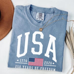May include: A light blue t-shirt with the word "USA" in large white letters. Below the word "USA" is the American flag, the years "1776" and "2026", and the text "250 YEARS OF FREEDOM". A gold necklace is draped across the shirt.