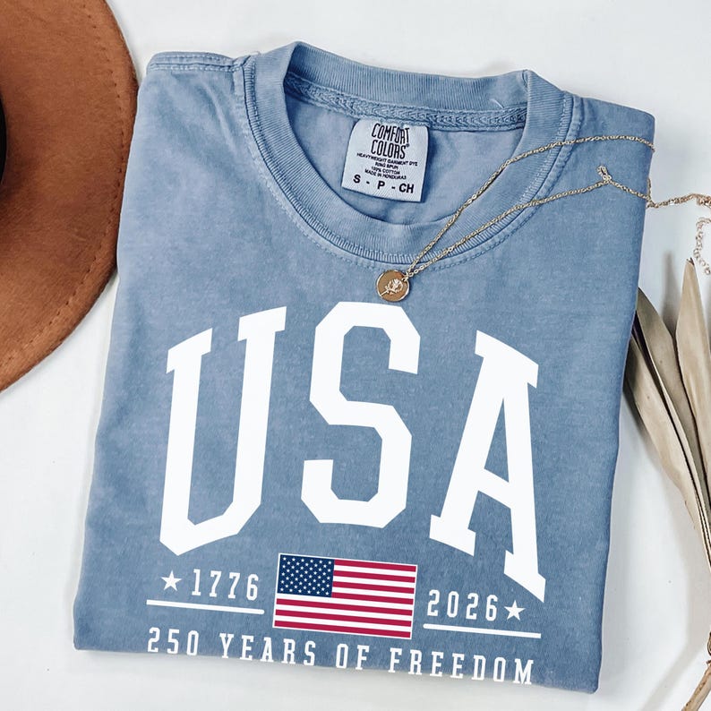 May include: A light blue t-shirt with the word "USA" in large white letters. Below the word "USA" is the American flag, the years "1776" and "2026", and the text "250 YEARS OF FREEDOM". A gold necklace is draped across the shirt.