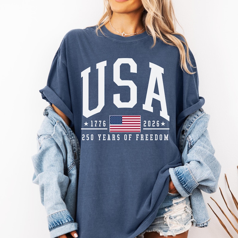 May include: Navy blue t-shirt with the word "USA" in white block letters. Below the word "USA" is a graphic of the American flag, the years "1776" and "2026", and the text "250 YEARS OF FREEDOM".
