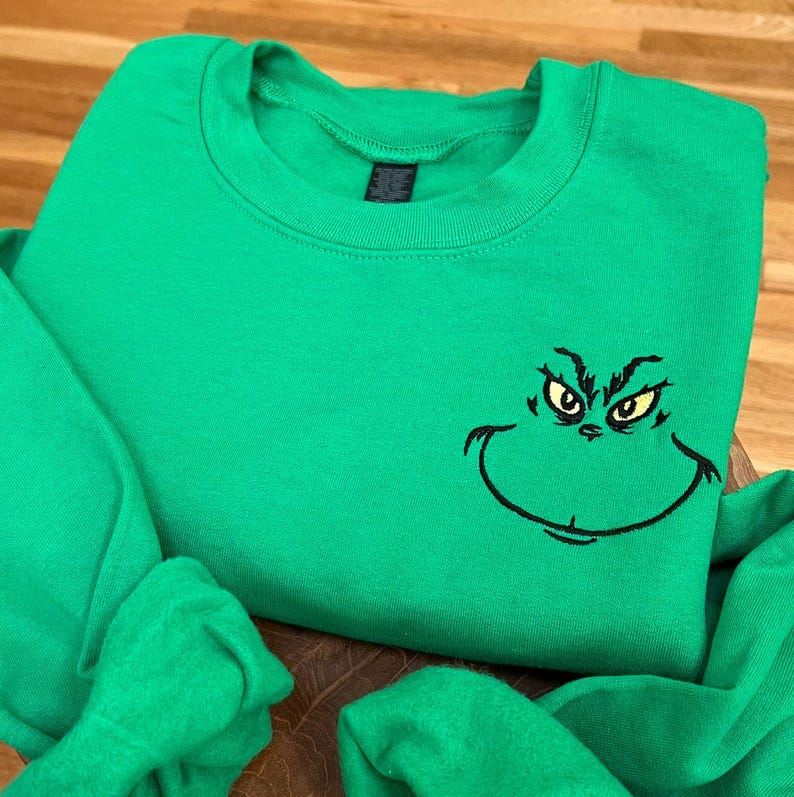 Puede incluir: Una sudadera verde brillante con una cara de Grinch bordada en negro. La sudadera tiene cuello redondo y mangas largas. La cara del Grinch incluye ojos amarillos y una sonrisa curva.