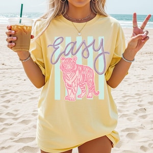 K&ouml;nnte beinhalten: Hellgelbes T-Shirt mit dem Wort "easy" in lila Schrift &uuml;ber einer rosa Tigerillustration. Der Tiger ist vor einem Hintergrund aus hellblauen vertikalen Streifen platziert. Das Shirt hat eine lockere, &uuml;bergro&szlig;e Passform.