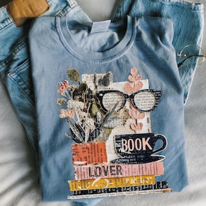 May include: A light blue t-shirt with a graphic design featuring a stack of books, a coffee cup, and eyeglasses. The design includes the word "BOOK LOVER" and floral elements. The shirt is paired with blue jeans and eyeglasses.