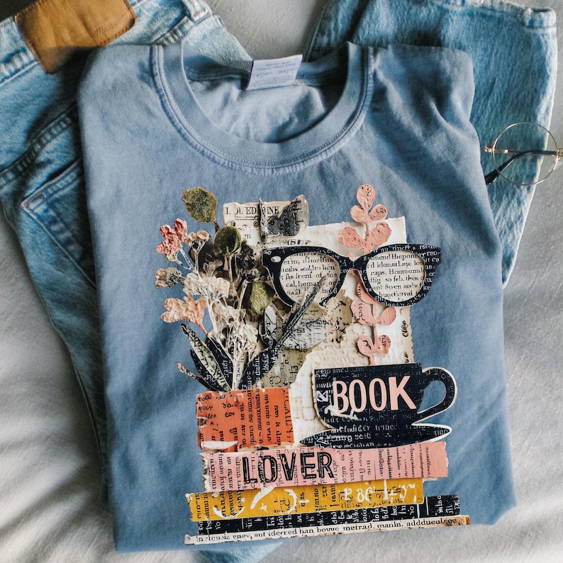 May include: A light blue t-shirt with a graphic design featuring a stack of books, a coffee cup, and eyeglasses. The design includes the word "BOOK LOVER" and floral elements. The shirt is paired with blue jeans and eyeglasses.