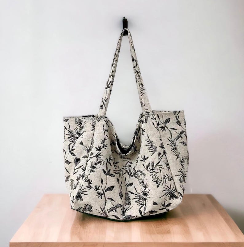 May include: A large, beige tote bag with black floral print. The bag has two long straps and is hanging from a hook.