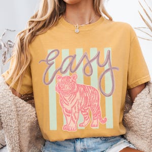 K&ouml;nnte beinhalten: Senfgelbes T-Shirt mit dem Wort "Easy" in Schreibschrift &uuml;ber einer rosa Tiger-Illustration. Der Tiger ist vor einem Hintergrund aus vertikalen hellgr&uuml;nen Streifen platziert. Das T-Shirt hat kurze &Auml;rmel.