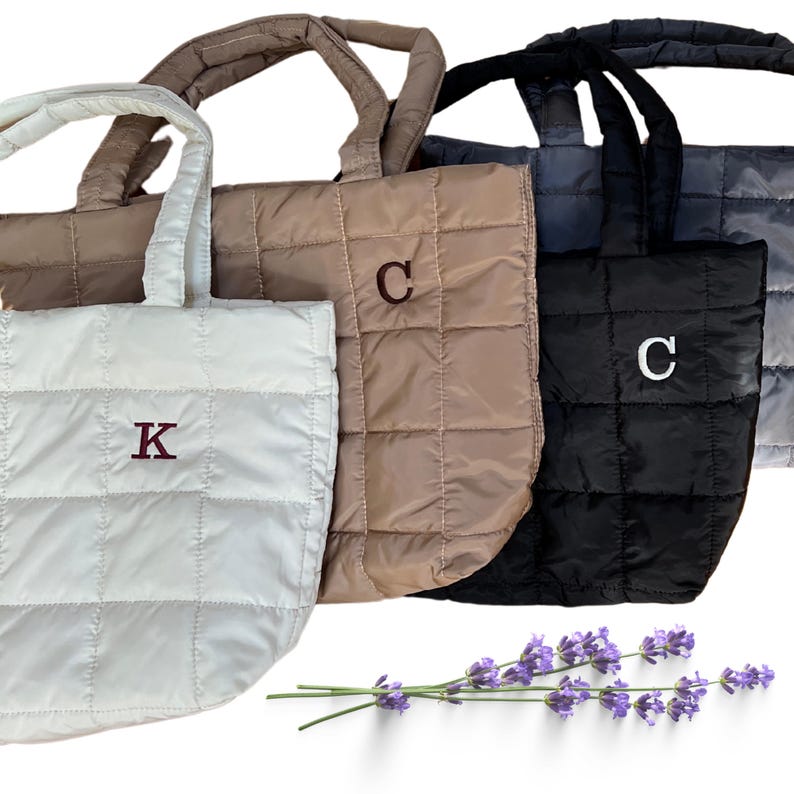 May include: Four quilted tote bags in shades of white, tan, brown, black, and gray. Each bag has a square pattern and a matching handle. Two bags have a single letter embroidered on them. A sprig of lavender is in the foreground.