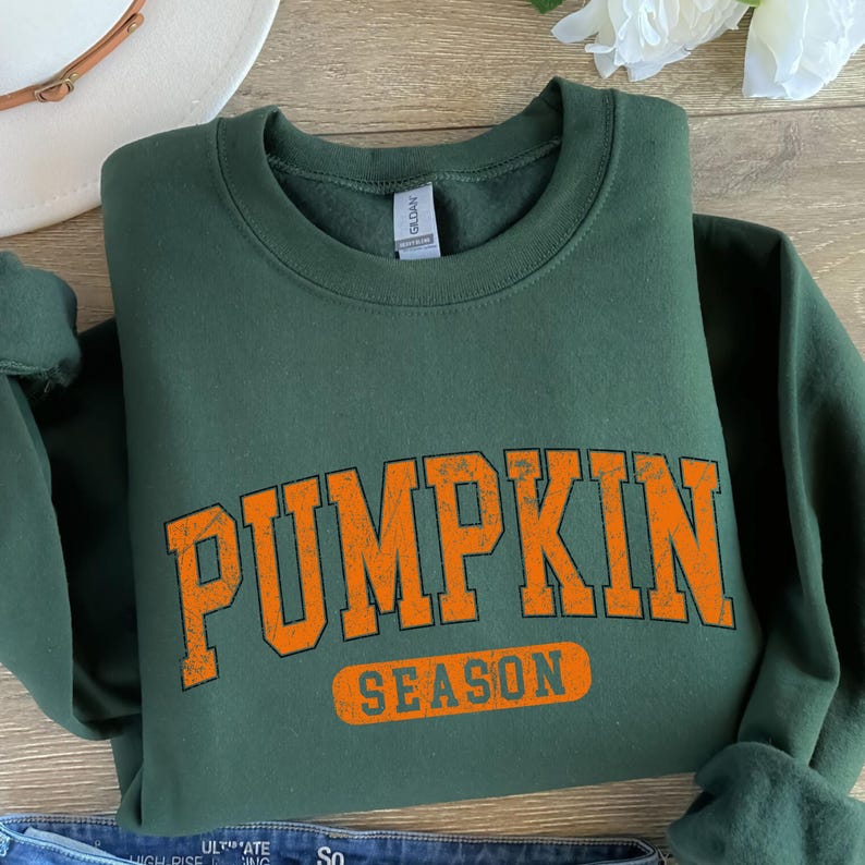 May include: A dark green sweatshirt with the words "PUMPKIN SEASON" printed in orange block letters. The sweatshirt is folded, with the text prominently displayed across the front. The design has a vintage, collegiate style.