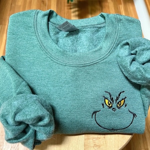 Puede incluir: Una sudadera verde con cuello redondo y mangas largas. La sudadera presenta una cara bordada del Grinch con ojos amarillos y un contorno negro. La prenda est&aacute; doblada sobre una superficie de madera.