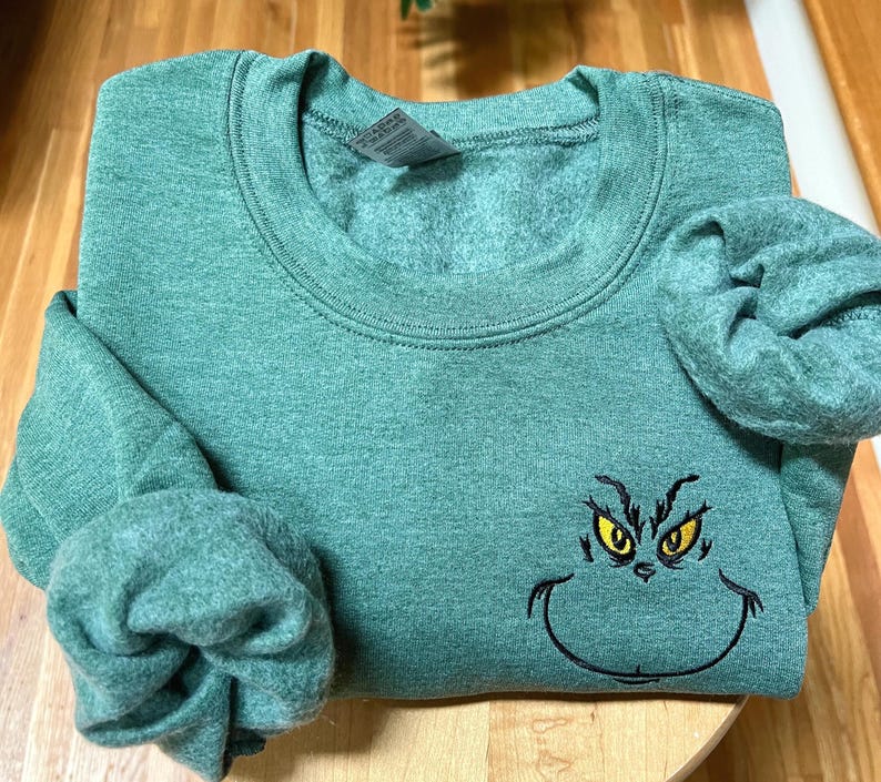 Puede incluir: Una sudadera verde con cuello redondo y mangas largas. La sudadera presenta una cara bordada del Grinch con ojos amarillos y un contorno negro. La prenda est&aacute; doblada sobre una superficie de madera.