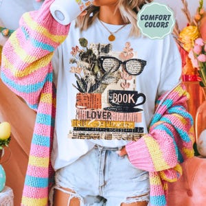 May include: White t-shirt with a graphic of a stack of books, a coffee cup, and glasses. The word "BOOK" is printed on the cup, and "LOVER" is printed on the books. The shirt is worn with a colorful striped sweater.