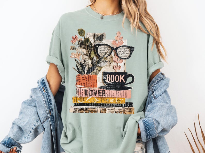 May include: A light green t-shirt featuring a collage design for book lovers. The design includes a stack of books, a coffee cup, eyeglasses, and floral elements. The word "BOOK" is printed on the cup, and "LOVER" is printed on the books.