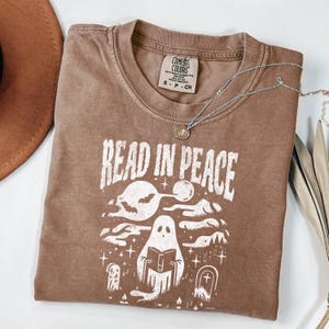 May include: A brown Comfort Colors t-shirt with the words "READ IN PEACE" in white. The graphic features a ghost reading a book, with bats, a moon, and tombstones. A silver necklace with a pendant is draped across the shirt.