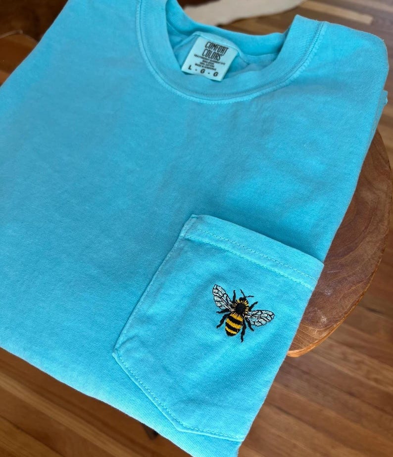 May include: A bright turquoise short-sleeve t-shirt featuring a single chest pocket. An embroidered honeybee with yellow and black stripes and white wings is centered on the pocket. The inner neck label reads Comfort Colors and size L G G.