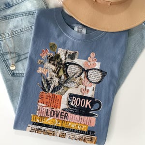May include: A blue Comfort Colors t-shirt featuring a collage design with the words "BOOK LOVER". The design includes a stack of books, eyeglasses, a coffee cup, and floral elements. The shirt is paired with blue jeans and a tan hat.