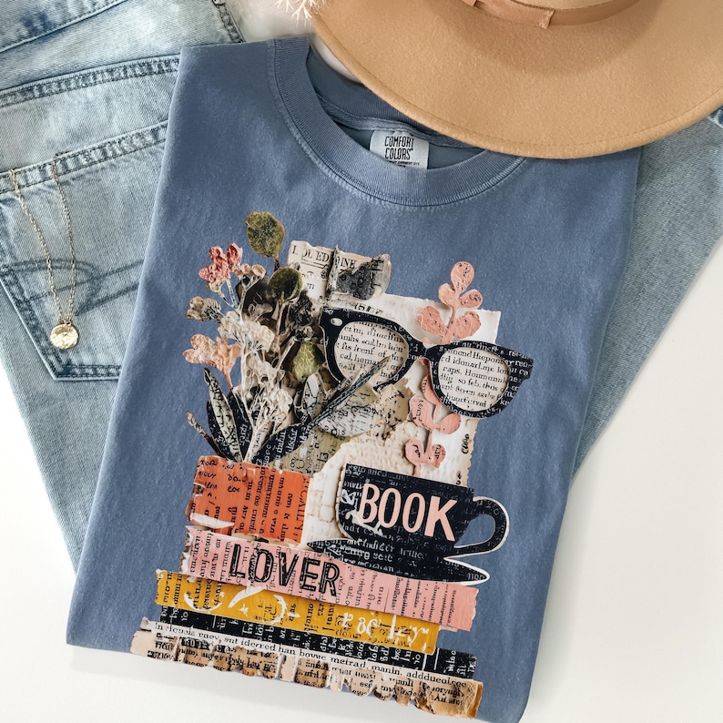 May include: A blue Comfort Colors t-shirt featuring a collage design with the words "BOOK LOVER". The design includes a stack of books, eyeglasses, a coffee cup, and floral elements. The shirt is paired with blue jeans and a tan hat.
