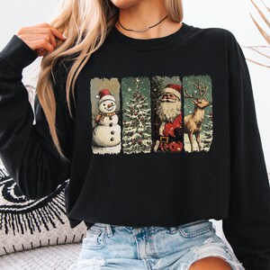 May include: Black long-sleeve sweatshirt featuring a festive Christmas design. The design includes a snowman, Christmas tree, Santa Claus, and a reindeer, each in a separate rectangular frame. The illustrations are in a vintage style with a snowy background.
