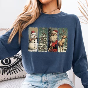May include: Navy blue sweatshirt featuring a festive design. The graphic includes a snowman, Christmas tree, Santa Claus, and a reindeer, all within vertical rectangular frames. The design has a vintage, slightly distressed look.
