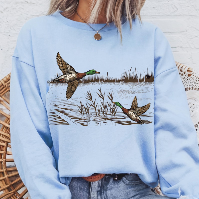 K&ouml;nnte beinhalten: Hellblaues Sweatshirt mit einer detaillierten Illustration von zwei Enten im Flug &uuml;ber Wasser mit Schilf. Die Enten haben braune, wei&szlig;e und gr&uuml;ne Markierungen. Das Kunstwerk ist ein Design zum Thema Natur.