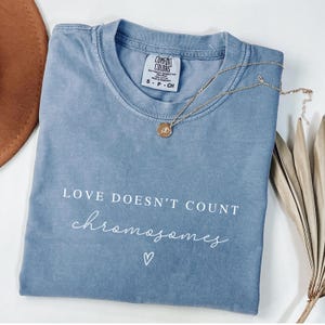 K&ouml;nnte beinhalten: Ein hellblaues T-Shirt mit dem Text "LOVE DOESN'T COUNT chromosomes" in wei&szlig;en Buchstaben. Ein kleines wei&szlig;es Herz befindet sich unter dem Text.