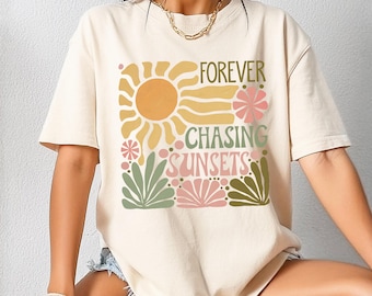 May include: Cream-colored t-shirt with the words "FOREVER CHASING SUNSETS" in a colorful, floral design. The graphic includes a yellow sun, pink flowers, and green foliage. The shirt is a casual, everyday wear item.