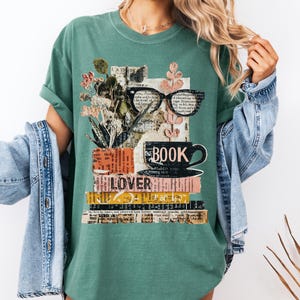 May include: Green t-shirt with a book-themed graphic. The design features a stack of books, a coffee cup, eyeglasses, and floral elements. The word "BOOK" is printed on the cup, and "LOVER" is visible on a book. The shirt is made of a soft material.