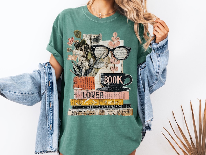 May include: Green t-shirt with a book-themed graphic. The design features a stack of books, a coffee cup, eyeglasses, and floral elements. The word "BOOK" is printed on the cup, and "LOVER" is visible on a book. The shirt is made of a soft material.
