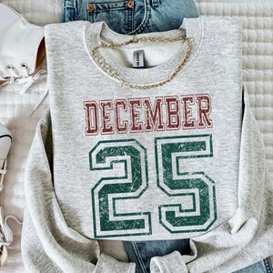 Pu&ograve; includere: Una felpa grigio melange con la scritta "DECEMBER 25" in lettere rosse e verdi sbiadite. Una collana a catena dorata &egrave; appoggiata sulla felpa. La felpa &egrave; abbinata a jeans blu e scarpe da ginnastica bianche.