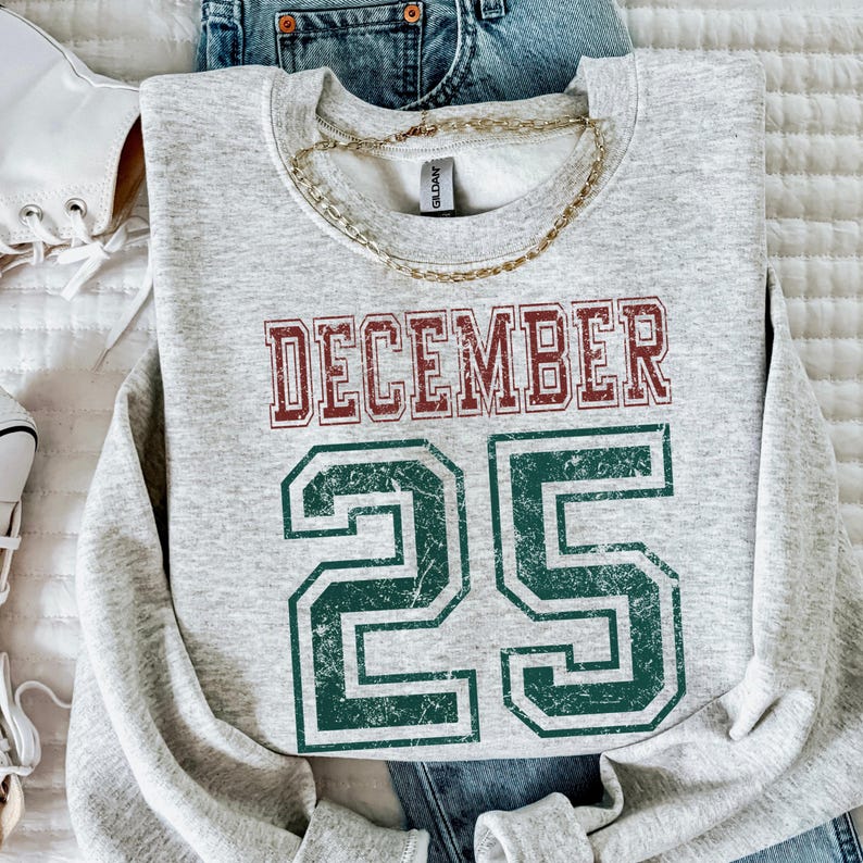 Pu&ograve; includere: Una felpa grigio melange con la scritta "DECEMBER 25" in lettere rosse e verdi sbiadite. Una collana a catena dorata &egrave; appoggiata sulla felpa. La felpa &egrave; abbinata a jeans blu e scarpe da ginnastica bianche.