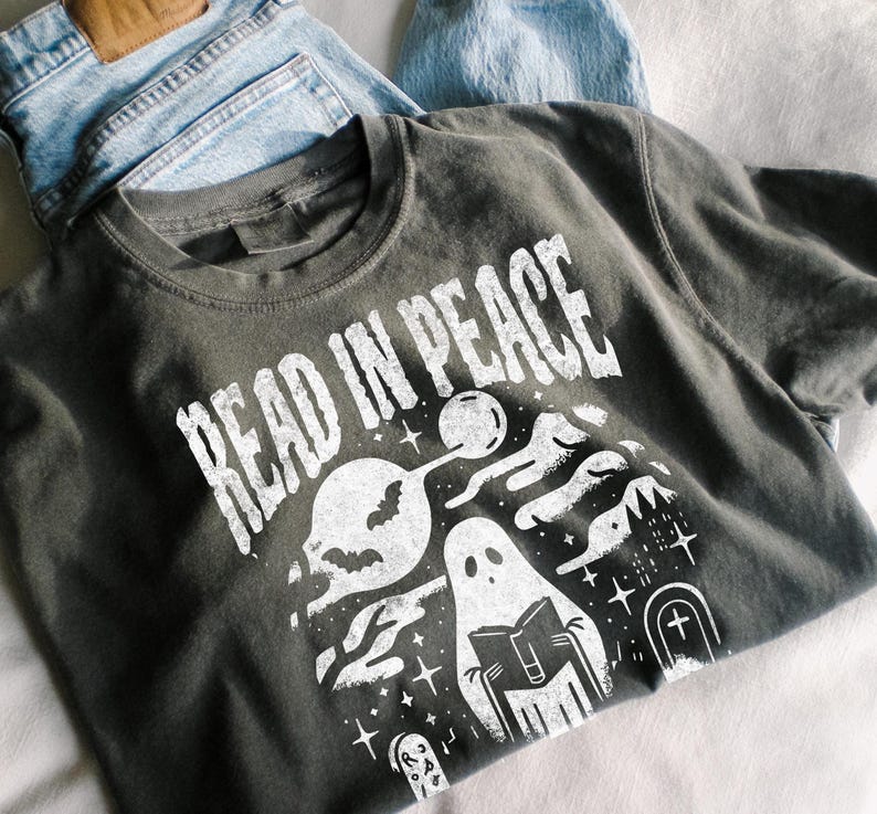 May include: Dark gray t-shirt with the words "READ IN PEACE" in white, featuring a spooky graphic of ghosts, a bat, and a graveyard scene. The shirt is folded on top of a pair of blue jeans.