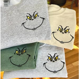 Puede incluir: Cuatro sudaderas en varios colores: gris, beige, verde y gris claro. Cada sudadera presenta una cara bordada con ojos amarillos y una sonrisa negra, que se asemeja a un personaje de dibujos animados. Las sudaderas est&aacute;n dobladas y apiladas.