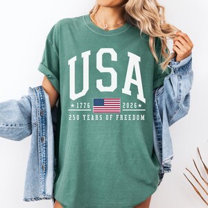 May include: A teal-colored t-shirt with the word "USA" in large white block letters. Below the word "USA" is the American flag, the years "1776" and "2026", and the text "250 YEARS OF FREEDOM".