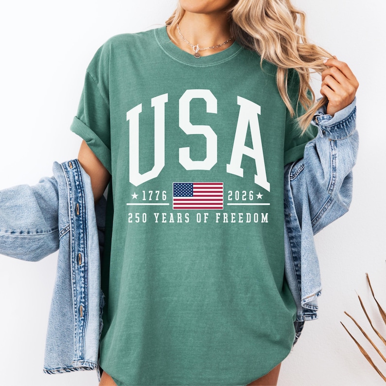 May include: A teal-colored t-shirt with the word "USA" in large white block letters. Below the word "USA" is the American flag, the years "1776" and "2026", and the text "250 YEARS OF FREEDOM".