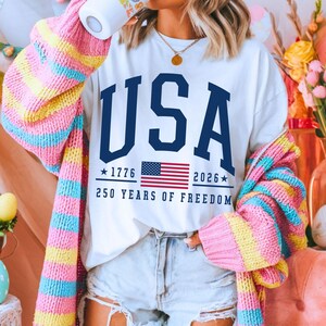 May include: White t-shirt with the word "USA" in navy blue, the American flag, and the text "1776 2026 250 Years of Freedom." The person is wearing a colorful striped sweater.