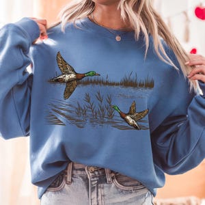 K&ouml;nnte beinhalten: Ein blaues Sweatshirt mit einer detaillierten Illustration von zwei Enten im Flug &uuml;ber Wasser und Schilf. Die Enten sind braun, gr&uuml;n und wei&szlig;. Das Sweatshirt hat eine einfarbige blaue Farbe.