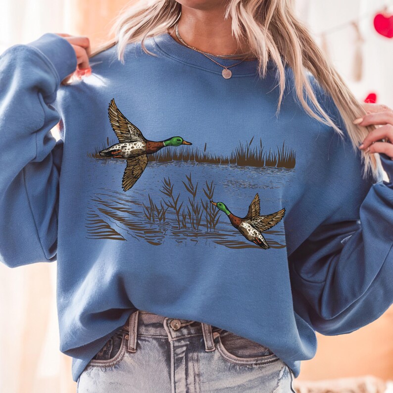 K&ouml;nnte beinhalten: Ein blaues Sweatshirt mit einer detaillierten Illustration von zwei Enten im Flug &uuml;ber Wasser und Schilf. Die Enten sind braun, gr&uuml;n und wei&szlig;. Das Sweatshirt hat eine einfarbige blaue Farbe.