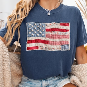 K&ouml;nnte beinhalten: Marineblaues T-Shirt mit einem Patchwork-Design der amerikanischen Flagge. Die Flagge verwendet rote, wei&szlig;e und blaue Stoffe mit Blumen- und Vichy-Mustern. Das T-Shirt hat einen Rundhalsausschnitt und kurze &Auml;rmel.