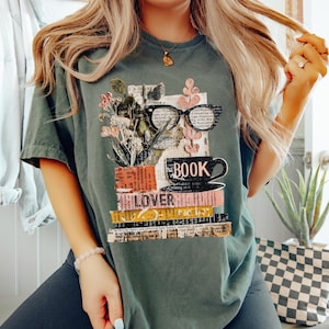 May include: A sage green t-shirt featuring a graphic design for book lovers. The design includes a stack of books, a coffee cup, eyeglasses, and the word "BOOK" and "LOVER". The design also includes floral elements.