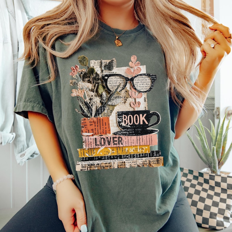 May include: A sage green t-shirt featuring a graphic design for book lovers. The design includes a stack of books, a coffee cup, eyeglasses, and the word "BOOK" and "LOVER". The design also includes floral elements.