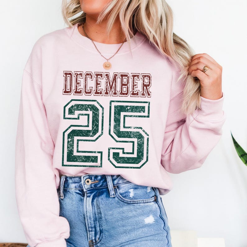 Pu&ograve; includere: Una felpa rosa chiaro con la scritta "DECEMBER 25" in un carattere in stile collegiale usurato. La parola "DECEMBER" &egrave; bordeaux e i numeri "25" sono verdi. La felpa &egrave; abbinata a jeans blu.